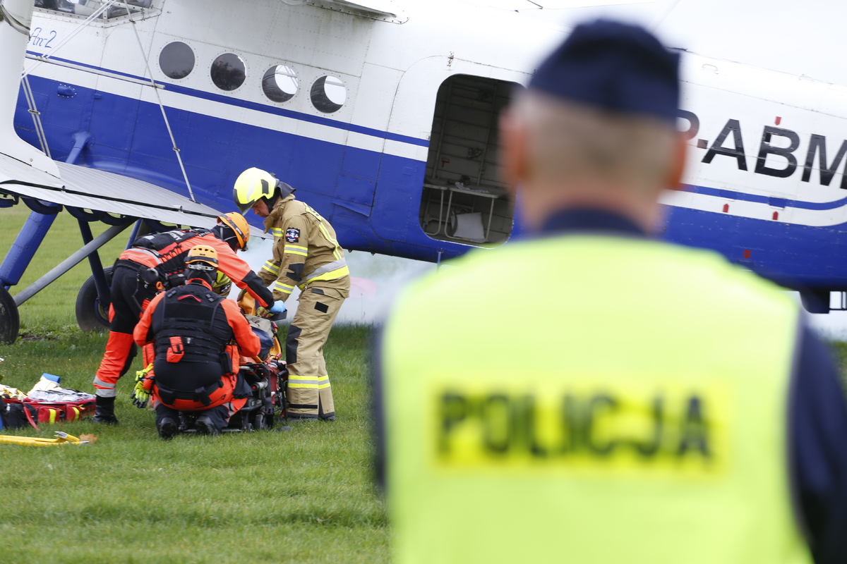 W pierwszym planie zdjęcia widzimy policjanta w mundurze w żółtej kamizelce z napisem policja. W głębi zdjęcia po lewej stronie strażacy i ratownicy medyczni udzielają pomocy pilotowi uszkodzonego samolotu.